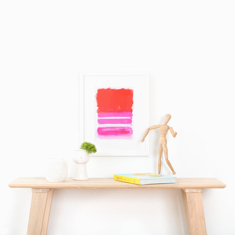 Red and pink abstract art in white frame above table with hat and plant